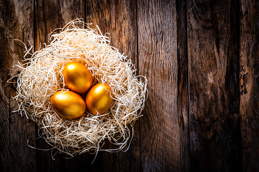 Holidays: overhead view of three gold colored Easter eggs arranged in straw nest at the left of an horizontal rustic wooden table leaving useful copy space for text and/or logo at the center. High resolution 42Mp studio digital capture taken with Sony A7rII and Sony FE 90mm f2.8 macro G OSS lens