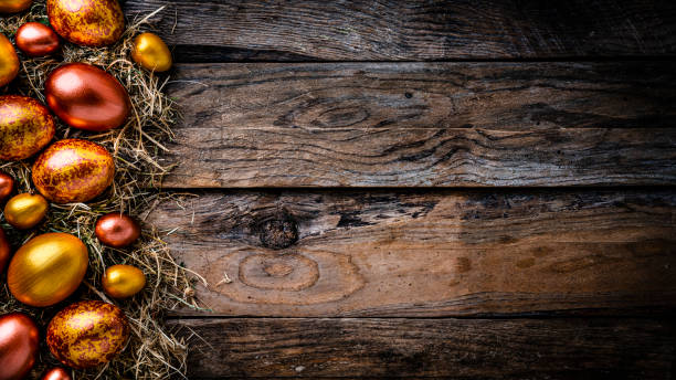 Holidays: overhead view of hand painted gold and bronze colored Easter eggs arranged in straw nest at the left of an horizontal rustic wooden table leaving useful copy space for text and/or logo at the center. High resolution 42Mp studio digital capture taken with Sony A7rII and Sony FE 90mm f2.8 macro G OSS lens