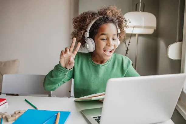 A little cute African American girl is on the sofa in the living room, she is using a laptop and waving to her teacher