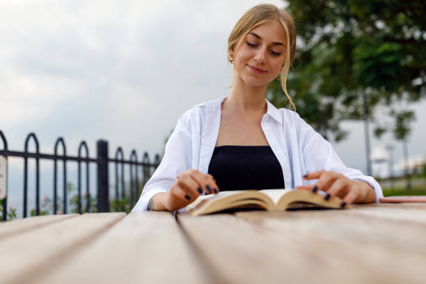 istock Young woman reading book at public park (Analog book reader) 1297658428