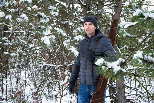 man in down jacket and jeans with shotgun in the case on the shoulder in the winter forest