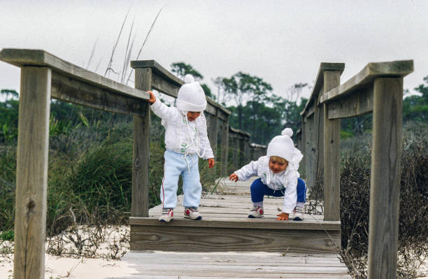 Two young toddlers are on a wooden boardwalk in sand dunes in the winter.  They are dressed in winter clothing. They're walking off the end of a wooden pier in the north Florida panhandle. There is no snow, but the weather is chilly. The girls are wearing knit hats and actively stepping down from the boardwalk.