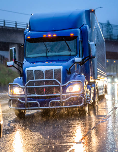 Powerful big rig blue semi truck with grille guard and turn on headlight with reefer semi trailer running on the wet highway road with reflection on a slippery surface in the rain Big rig stylish industrial blue semi truck with turned on headlights transporting cargo in refrigerator semi trailer running on the twilight wet road with light reflection surface in rain weather truck rain stock pictures, royalty-free photos & images