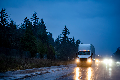 Big rig industrial white semi truck with turned on headlights transporting cargo in dry van semi trailer running on the night dark twilight wet road with reflection on surface in rain weather