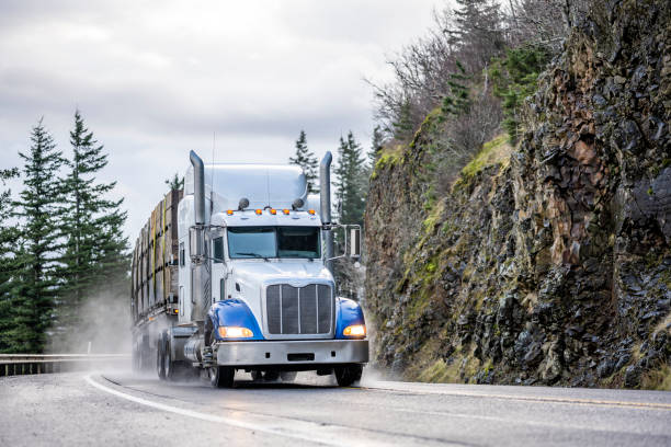 istock Powerful big rig white semi truck tractor transporting cargo in boxes stacked on flat bed semi trailer running on winding mountain road in Columbia Gorge at rainy weather 1297668906
