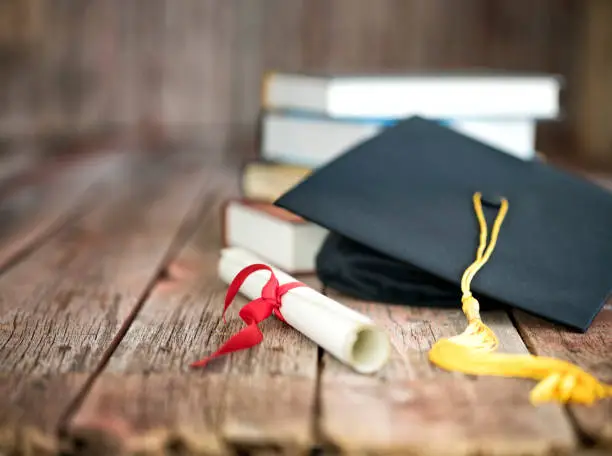 Graduation cap and diploma with a stack of books on a wood background