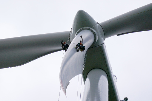 View from bottom on wind-turbine and two rope access technicians, industrial climbers rappelling down from hub on blade on the ropes, doing inspection, blade repair and waving on camera