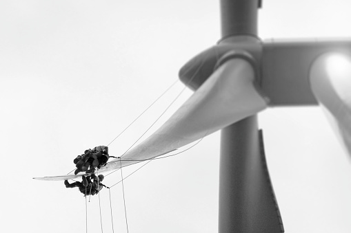 Two Professional Rope access technicians -industrial climber working and cooperating on tip of blade of wind turbine. Hanging in the ropes and repairing edge, doing inspection and repairing serrations and fibber glass. They are directly above