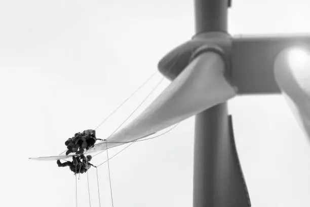 Photo of Two Professional Rope access technicians -industrial climber working and cooperating on tip of blade of wind turbine. Hanging in the ropes and repairing edge, doing inspection and repairing serrations and fibber glass. They are directly above