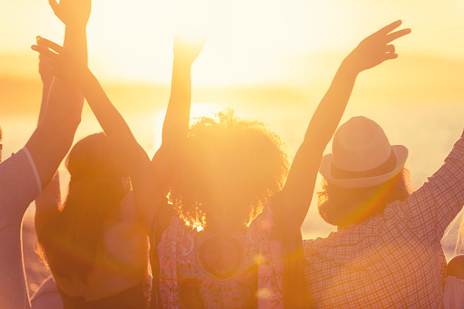 Group of friends sitting on the beach at sunset/sunrise. They are watching the sun set over the ocean. They are celebrating with their arms in the air. One woman is making a peace sign. Backlit silhouette with copy space.