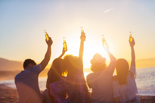 Group of young people partying on the beach at sunset. They are all holding bottles of beer and celebrating with a toast. Silhouette with the beach, sun and sea in the background.