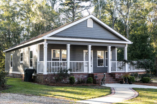 Close up of small blue gray mobile home with a front and side porch with white railing Small blue gray mobile home with a front and side porch. It has a white porch railing out in the country on land shared by a larger siding house. house stock pictures, royalty-free photos & images
