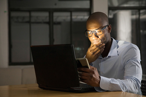 Mature black man while working on his laptop and mobile phone at night. He looks pensively at what he sees on his screens.