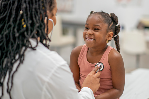 A super adorable girl of African ethnicity is at her doctor's office for a check up. She is sitting on the examination table while her doctor is using a stethoscope to listen to her chest.