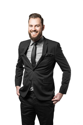 Handsome and laughing bearded businessman wearing a black suit and tie looking positive with both hands in his pockets. White background.