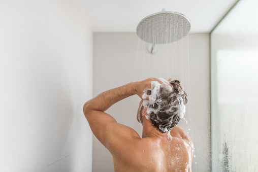 Hombre tomando una ducha lavando el cabello bajo el agua cayendo de la ducha de lluvia en el baño de lujo a ras de suelo. Duchando a la persona joven en el estilo de vida del hogar. Rutina matutina de cuidado corporal a la luz del sol photo