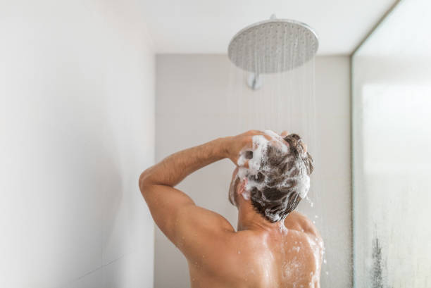 Man taking a shower washing hair under water falling from rain showerhead in luxury walk-in bath. Showering young person at home lifestyle. Body care morning routine in sunlight stock photo