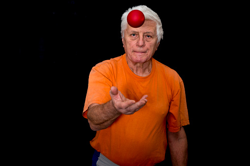 Front view of Senior man, a Parkinson Disease patient, who is standing  in front of monitor, at home with black background. He juggles with a little ball in the framework of therapeutic exercises conducted over the Internet by dancing  therapist for Parkinson Disease. Online is only possible therapy as it is Covid-19 lockdown time.