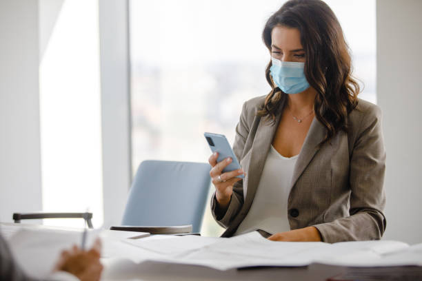 istock Businesswoman with protective face mask sitting at conference table and using phone 1297722767