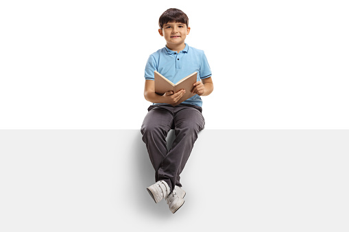 Cute boy sitting on a blank panel with a book in his hands isolated on white background