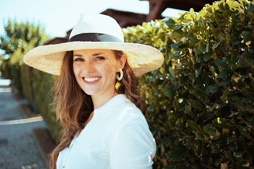 Portrait of smiling stylish middle aged housewife in white shirt with hat outdoors near green wall.