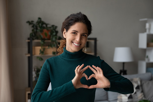 Portrait of smiling millennial Caucasian woman show heart love hand gesture or sign. Happy young female look at camera demonstrate support and care, feel supportive. Volunteer, charity concept.