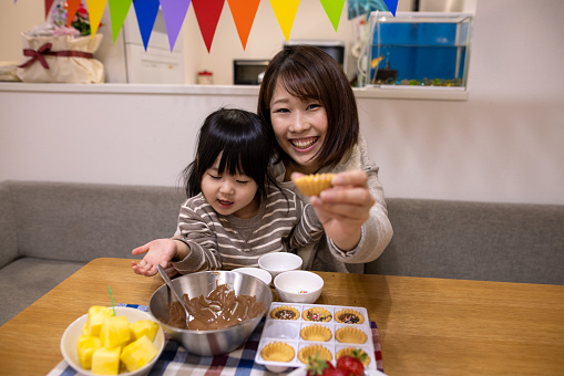 Young mother celebrating her baby boy's first birthday with her little daughter at home. Making small tarts and eating with fruits, sending birthday presents, singing birthday song, etc.