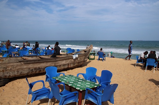 Lomé, Togo - June 23, 2019: Restaurant with blue chairs at the public beach of Lomé, the capital of Togo, West Africa. Guests have a drink. Fishing boats are between the tables.