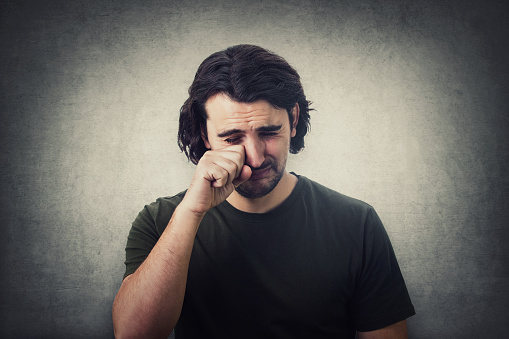 Portrait of desperate young man crying and wiping the tears with his hand isolated on grey wall background. Dissatisfied and disappointed guy sobbing and weeping to camera.