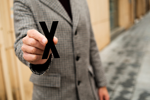 closeup of a young caucasian person standing on the street, wearing a coat, showing an X, for the third gender category
