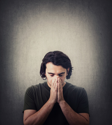 Portrait of depressed young man keeps hands together as prayer, eyes closed, head down isolated on grey background. Desperate guy praying for help. Concept of meditation, calming down and inner peace