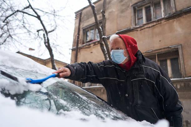 aktywny starszy mężczyzna, usuwając śnieg z przedniej szyby samochodu z plastikowym skrobakem lodu i miotłą podczas noszenia ochronnej maski na twarz z powodu wybuchu covid-19 - snow car window ice scraper zdjęcia i obrazy z banku zdjęć