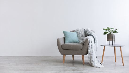 Minimalist interior of bedroom, cabinet or living room. Gray modern armchair with blue pillow and soft blanket, on table pot with plant, home design with light wall in scandinavian style, panorama