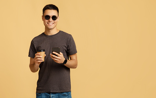 Stylish male model on break with gadget. Cheerful millennial handsome man student in sunglasses holds cup of coffee takeaway and smartphone, isolated on beige background, studio shot, free space