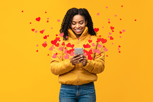 Happy African Woman Using Phone Sending Hearts And Like Icons Texting On Valentine's Holiday In Online Dating App Standing Over Yellow Studio Background. Love Chat, Romance And Networking