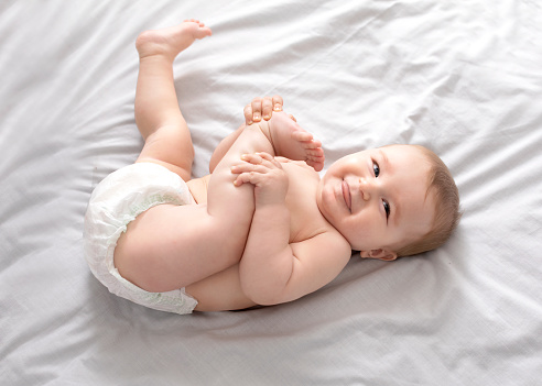 Beautiful caucasian baby laying on bed, playing with feet and smiling at camera, top view, copy space. Adorable toddler in diaper having fun alone, lying on white, laughing. Toddler, baby, infant