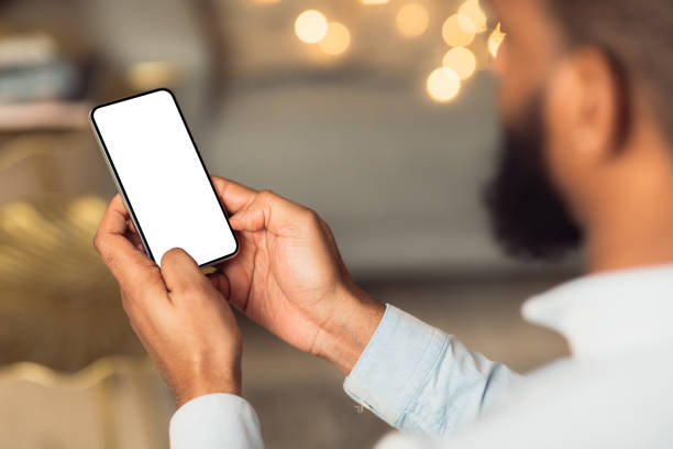 Black man holding phone with empty mockup screen People And Technology Concept. Over The Shoulder View Of Unrecognizable African American Man Using Smart Phone With White Empty Screen For Mockup, Sitting At Home, Making Video Call Or Chatting man looking at phone over shoulder stock pictures, royalty-free photos & images