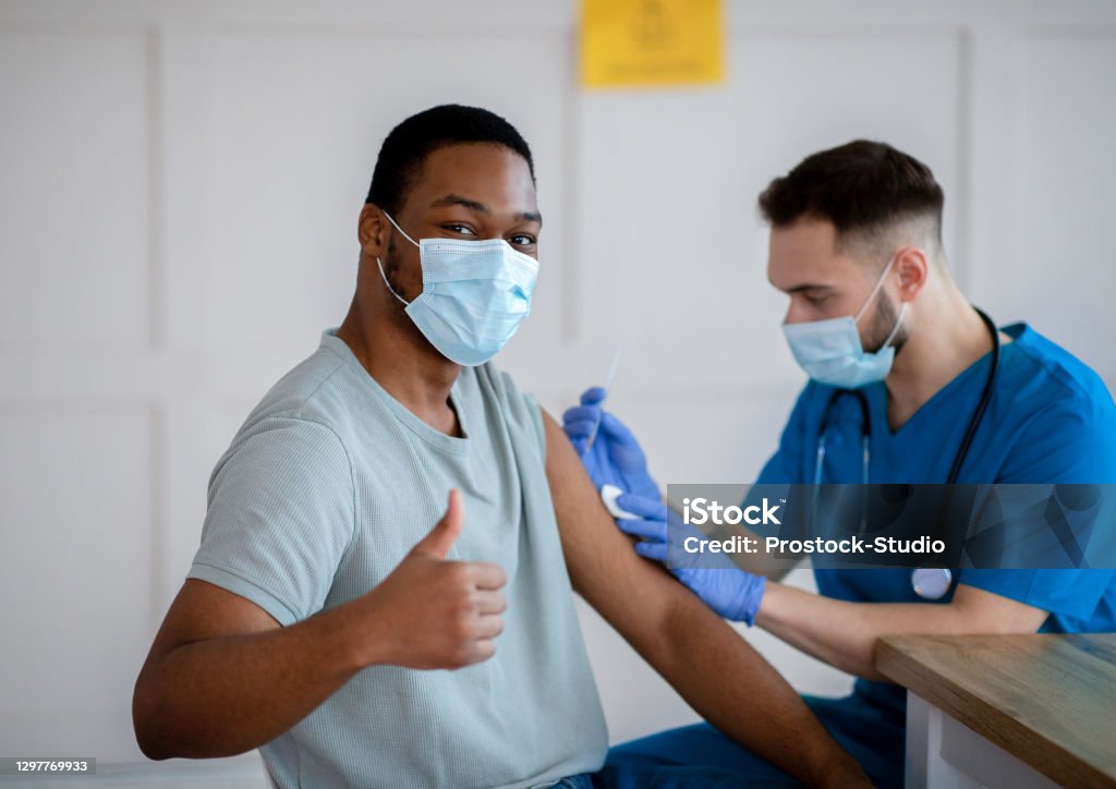 African American man in mask gesturing thumb up during coronavirus vaccination, approving of covid-19 immunization African American man in antiviral mask gesturing thumb up during coronavirus vaccination, approving of covid-19 immunization. Young doctor giving vaccine injection to male patient COVID-19 Vaccine Stock Photo