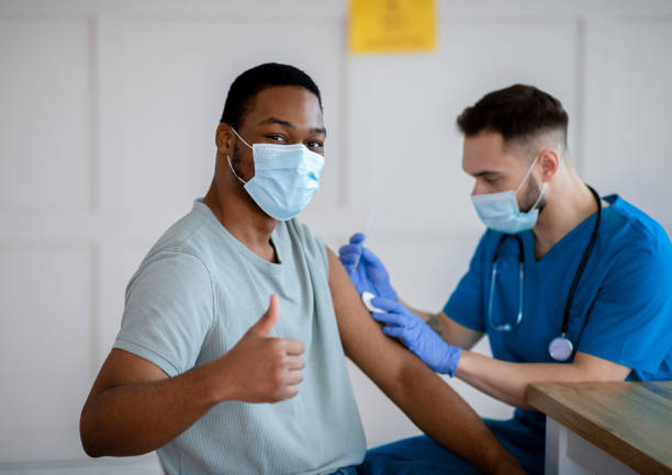 African American man in mask gesturing thumb up during coronavirus vaccination, approving of covid-19 immunization African American man in antiviral mask gesturing thumb up during coronavirus vaccination, approving of covid-19 immunization. Young doctor giving vaccine injection to male patient pneumonia photos stock pictures, royalty-free photos & images