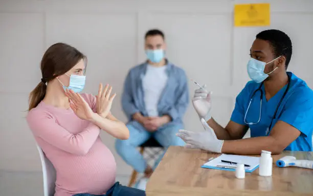 Photo of Pregnant woman in face mask scared to make coronavirus vaccine injection, making STOP gesture at clinic, panorama