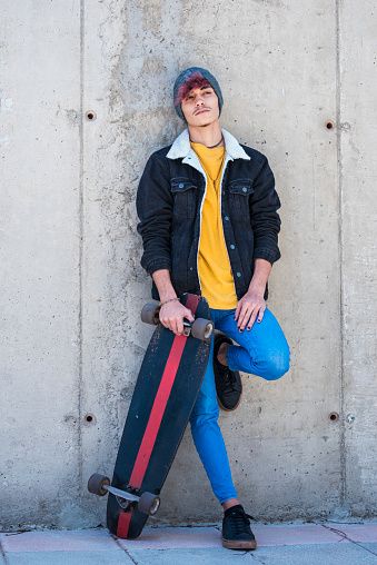 Portrait of young and alternative boy standing with skate board and colourful clothes - modern people and youthful lifestyle - cement wall in background for city concept