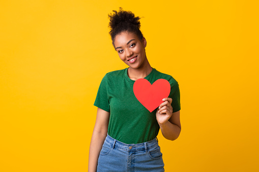 Love Is In The Air. Portrait of smiling young african american woman holding red paper heart, posing and looking at camera isolated on yellow studio background. Valentine's day, bonding, romance