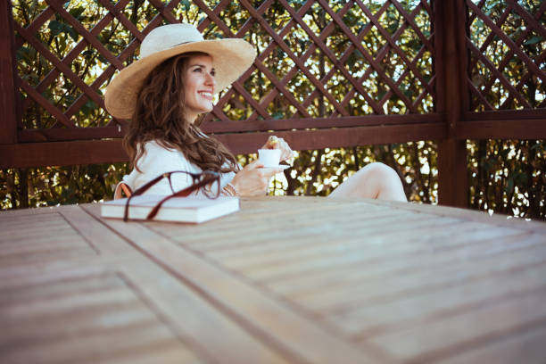 smiling stylish woman in white shirt with book, hat and eyeglasses sitting at the table drinking coffee in the terrace of guest house hotel.