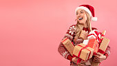 Happy millennial lady in Santa hat and Christmas sweater holding pile of gift boxes on pink background, banner design