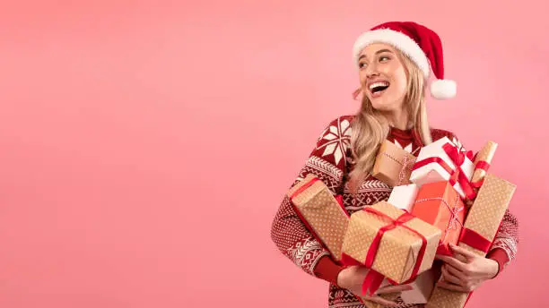 Photo of Happy millennial lady in Santa hat and Christmas sweater holding pile of gift boxes on pink background, banner design