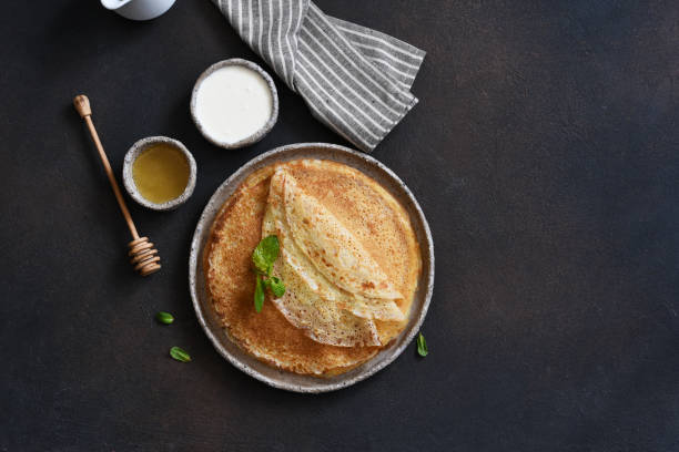istock Thin pancakes with sour cream and honey on a dark concrete background. A stack of pancakes. View from above. 1297776301