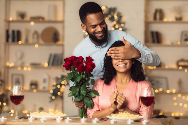 Young black man giving flowers to woman, covering eyes Greeting With Holidays. Smiling black man covering his woman eyes and giving her bunch of red roses, making surprise to beautiful lady. African american couple celebrating together at home or cafe boyfriend stock pictures, royalty-free photos & images