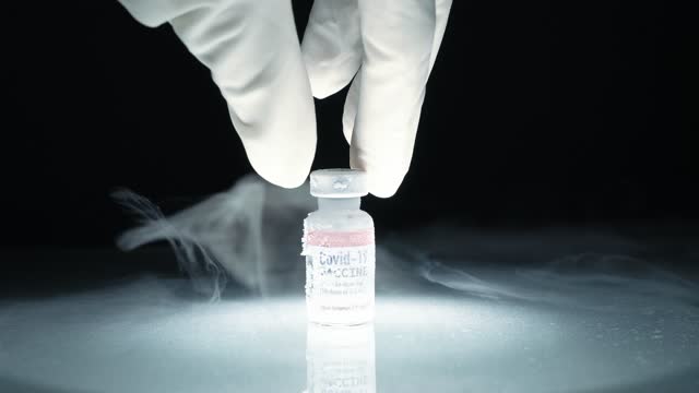 Close up of researcher's hand pick up frozen cold Covid-19 vaccine vial stored in subzero low temperature -70 celsius with flowing fog in dark background.