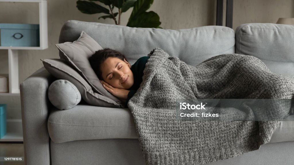 Happy young woman sleeping under blanket on sofa Happy millennial Caucasian woman lying under blanket on cozy couch at home sleeping or dreaming. Smiling calm young female relax rest on sofa in living room, relieve negative emotions daydreaming. Napping Stock Photo
