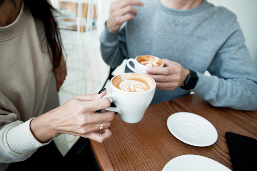 Young couple drinking hot beverages. Close up woman and man sitting in cafe, holding warm cups of coffee on table. Man and woman spending weekend in cozy coffeehouse together. Romantic date concept